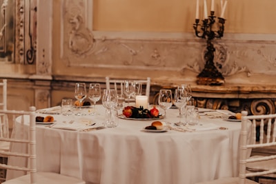 The image features an elegantly set dining table adorned with glassware, cutlery, dishes with bread rolls, and a decorative centerpiece with red fruits and a white candle. The chairs surrounding the table are white, complementing the tablecloth's color. In the background, intricate wall embellishments and a vintage candle holder add a sophisticated and classic touch to the atmosphere.