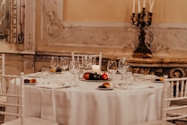 The image features an elegantly set dining table adorned with glassware, cutlery, dishes with bread rolls, and a decorative centerpiece with red fruits and a white candle. The chairs surrounding the table are white, complementing the tablecloth's color. In the background, intricate wall embellishments and a vintage candle holder add a sophisticated and classic touch to the atmosphere.