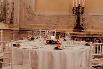 The image features an elegantly set dining table adorned with glassware, cutlery, dishes with bread rolls, and a decorative centerpiece with red fruits and a white candle. The chairs surrounding the table are white, complementing the tablecloth's color. In the background, intricate wall embellishments and a vintage candle holder add a sophisticated and classic touch to the atmosphere.