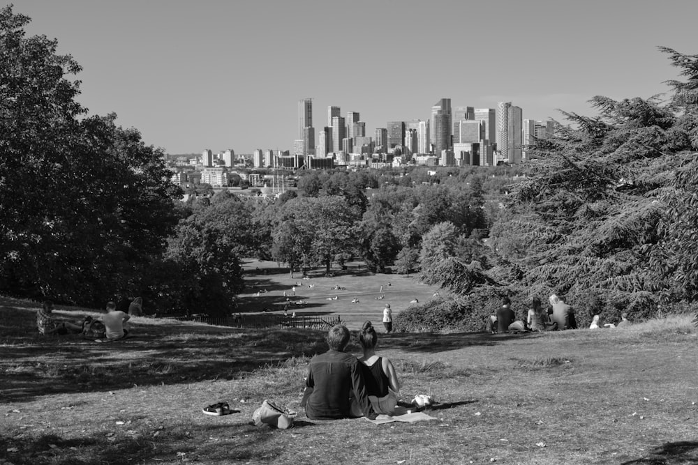 grayscale photo of people sitting on grass field near city buildings