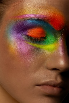 A close-up of a person's face with eyes closed, showcasing bold and vibrant multicolored makeup around the eyes. The makeup is skillfully blended, featuring hues of red, orange, yellow, green, blue, and purple, creating a striking and artistic appearance.