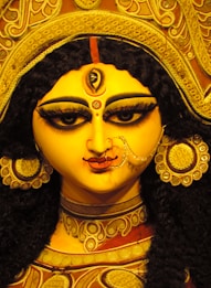 A vibrant, detailed idol of a goddess with striking features. The face is adorned with ornate jewelry and an elaborate headpiece. The eyes are heavily outlined, with a third eye on the forehead, symbolizing wisdom. The heirloom nose ring and large earrings enhance the divine appearance.
