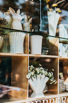 Ceramic vases, some with striped patterns, and decorative bunny figures are arranged on wooden shelves. A clear glass countertop supports other items, including a white vase filled with small white and yellow flowers. Reflection from the glass creates an overlay of natural surroundings.