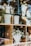 Ceramic vases, some with striped patterns, and decorative bunny figures are arranged on wooden shelves. A clear glass countertop supports other items, including a white vase filled with small white and yellow flowers. Reflection from the glass creates an overlay of natural surroundings.