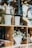 Ceramic vases, some with striped patterns, and decorative bunny figures are arranged on wooden shelves. A clear glass countertop supports other items, including a white vase filled with small white and yellow flowers. Reflection from the glass creates an overlay of natural surroundings.