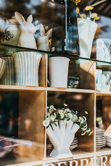 Ceramic vases, some with striped patterns, and decorative bunny figures are arranged on wooden shelves. A clear glass countertop supports other items, including a white vase filled with small white and yellow flowers. Reflection from the glass creates an overlay of natural surroundings.