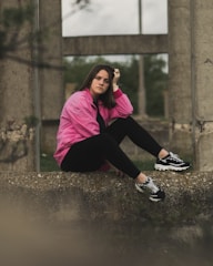A person wearing a pink jacket and black leggings sits thoughtfully on a concrete ledge in an outdoor setting with industrial concrete structures in the background.