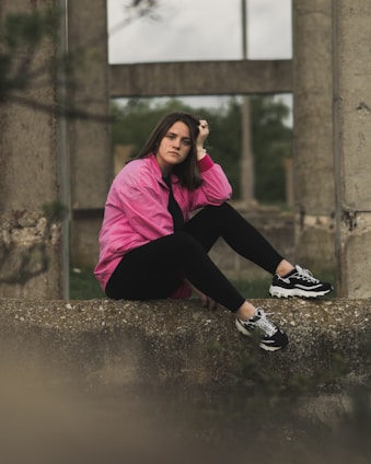 A person wearing a pink jacket and black leggings sits thoughtfully on a concrete ledge in an outdoor setting with industrial concrete structures in the background.