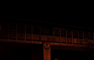 A dimly lit bridge or elevated structure with a series of vertical bars and a circular sign featuring the letters 'T E K'. The overall scene is dark with minimal ambient light, suggesting a nighttime setting. There is also some foliage visible in the lower right corner.