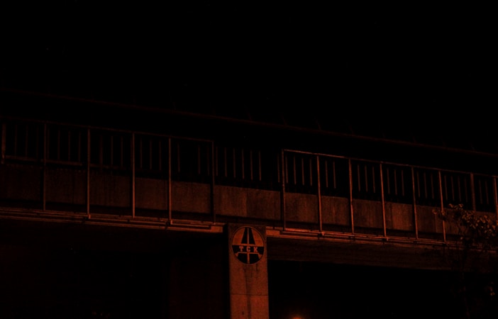 A dimly lit bridge or elevated structure with a series of vertical bars and a circular sign featuring the letters 'T E K'. The overall scene is dark with minimal ambient light, suggesting a nighttime setting. There is also some foliage visible in the lower right corner.