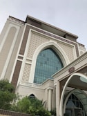 A modern building with Islamic architectural elements, featuring a large arched window with a grid of glass panes. The facade includes intricate geometric patterns and ornamental details in a combination of cream and light brown colors. The prominent signage at the top reads 'Crowne Plaza,' indicating it is a hotel. Lush green vegetation is present in the foreground.