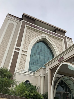 A modern building with Islamic architectural elements, featuring a large arched window with a grid of glass panes. The facade includes intricate geometric patterns and ornamental details in a combination of cream and light brown colors. The prominent signage at the top reads 'Crowne Plaza,' indicating it is a hotel. Lush green vegetation is present in the foreground.