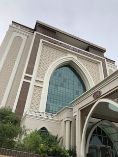 A modern building with Islamic architectural elements, featuring a large arched window with a grid of glass panes. The facade includes intricate geometric patterns and ornamental details in a combination of cream and light brown colors. The prominent signage at the top reads 'Crowne Plaza,' indicating it is a hotel. Lush green vegetation is present in the foreground.