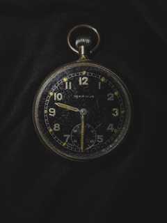 A vintage pocket watch with a black face and gold numbers and hands, showing signs of wear and scratches. It is placed against a dark fabric background, emphasizing its classic design and aged appearance. The watch features a smaller second dial at the bottom and a crown at the top for winding.