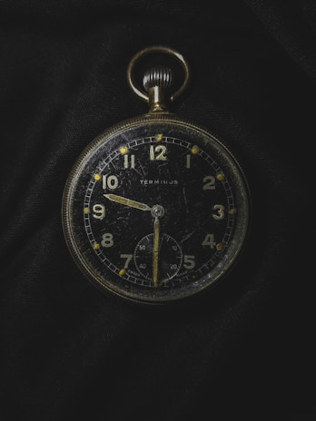 A vintage pocket watch with a black face and gold numbers and hands, showing signs of wear and scratches. It is placed against a dark fabric background, emphasizing its classic design and aged appearance. The watch features a smaller second dial at the bottom and a crown at the top for winding.