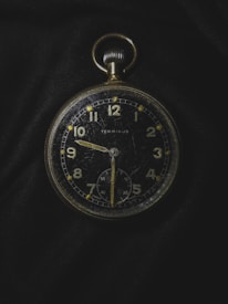 A vintage pocket watch with a black face and gold numbers and hands, showing signs of wear and scratches. It is placed against a dark fabric background, emphasizing its classic design and aged appearance. The watch features a smaller second dial at the bottom and a crown at the top for winding.
