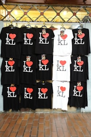 A display of multiple t-shirts with the text 'I ❤️ KL', arranged in two rows. Most shirts are black with white and red print, while a couple are white with black and red print. The shirts are hung neatly on a wire frame.