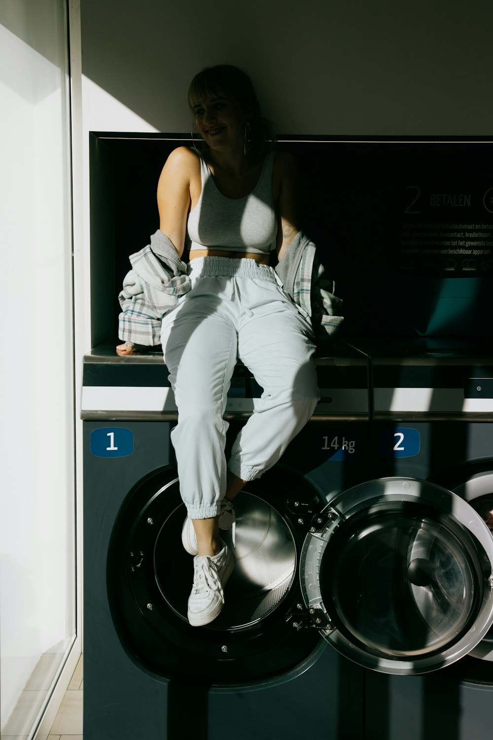 woman in white tank top and gray pants leaning on front load washing machine