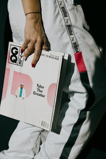 A person wearing a white outfit with black stripes is holding a GQ magazine featuring Tyler, The Creator. The magazine cover is predominantly white with pink circular graphics.