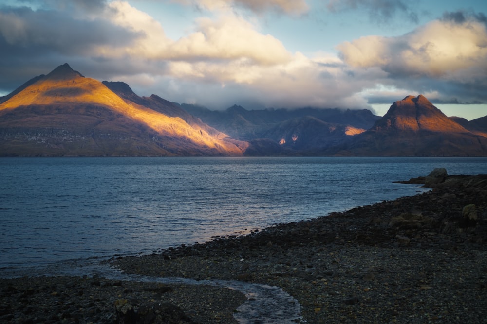 body of water near mountain under white clouds during daytime