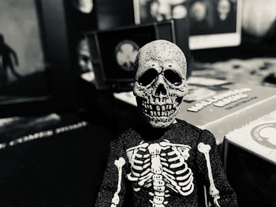 A small skeleton figurine is positioned in the foreground, with a skeletal body suit and a detailed skull. The background displays various game boxes and blurry, indistinct images, contributing to a mysterious and eerie atmosphere.