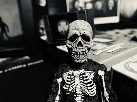 A small skeleton figurine is positioned in the foreground, with a skeletal body suit and a detailed skull. The background displays various game boxes and blurry, indistinct images, contributing to a mysterious and eerie atmosphere.