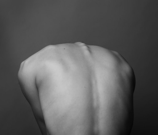 A monochrome photograph depicting the bare back of a person. The image focuses on the smooth contours and natural form of the human back, set against a plain, dark background.