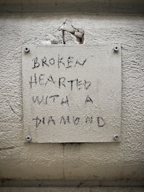 A textured wall features a pale rectangular plaque with an inscription that reads 'BROKEN HEARTED WITH A DIAMOND' in a rough, handwritten style. The wall has visible cracks and dirt, suggesting age or wear.