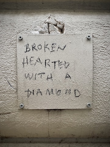 A textured wall features a pale rectangular plaque with an inscription that reads 'BROKEN HEARTED WITH A DIAMOND' in a rough, handwritten style. The wall has visible cracks and dirt, suggesting age or wear.