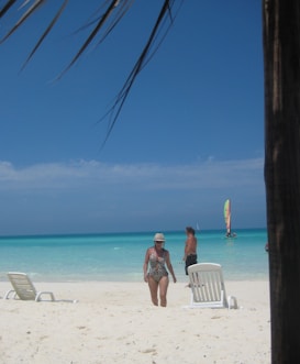 A sandy beach with clear blue water and a sunny sky. A woman in a patterned swimsuit and hat walks near two white lounge chairs. In the background, a man stands near the water, and a colorful sailboat is visible on the ocean. Palm fronds partially shadow the scene from above.