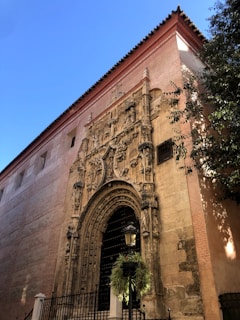 An ornate, historic architectural facade features intricate carvings and sculptures surrounding a large arched door. The building is made of light brown stone, with a tall wall extending upward. A black iron fence and a lantern with greenery are present in the foreground. Sunlight casts dynamic shadows, emphasizing the detailed craftsmanship.