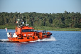 A bright orange pilot boat navigates through calm waters, leaving a trail of white waves behind. The boat carries several flags on its mast and is marked with the word 'PILOT' on its side. In the background, lush green trees line the shore under a clear blue sky.