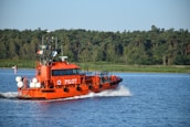 A bright orange pilot boat navigates through calm waters, leaving a trail of white waves behind. The boat carries several flags on its mast and is marked with the word 'PILOT' on its side. In the background, lush green trees line the shore under a clear blue sky.