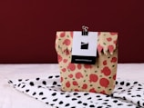 A beige paper bag adorned with a red pomegranate pattern is placed on a surface covered with a black and white pomegranate patterned cloth. The bag is held closed at the top with a black binder clip attached to a card.