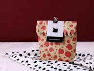 A beige paper bag adorned with a red pomegranate pattern is placed on a surface covered with a black and white pomegranate patterned cloth. The bag is held closed at the top with a black binder clip attached to a card.