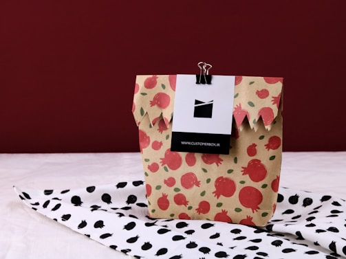 A beige paper bag adorned with a red pomegranate pattern is placed on a surface covered with a black and white pomegranate patterned cloth. The bag is held closed at the top with a black binder clip attached to a card.