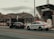 A police officer stands beside a black sedan that is pulled over next to the sidewalk. A police car with its lights on is parked behind the sedan. The scene is set near a dock, with a large ship labeled 'Hinch Spirit' in the background. There's a mountainous landscape under an overcast sky.