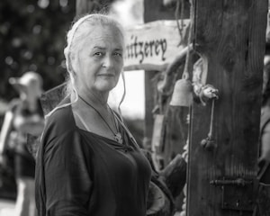 An older woman with white hair, wearing a dark shirt and necklace, standing outdoors. She is next to a wooden structure decorated with rustic items, including a bell. The background is softly blurred, with another person visible. The image is predominantly black and white, except for subtle hints of color.