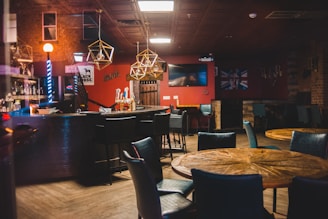 A cozy bar with a wooden floor and modern decor is minimally lit with ambient lighting. The bar counter is equipped with bar stools, liquor bottles, and neon lights displaying various signs. Geometric pendant lights hang overhead, illuminating the space. Wooden tables and upholstered chairs are arranged for seating, and a large screen is mounted on the wall.