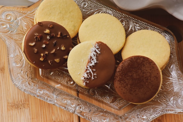 An assortment of cookies is displayed on an ornate, clear glass tray. There are six round cookies, three with toppings and three plain. The topped cookies have chocolate coatings with various garnishes including chopped nuts, shredded coconut, and a dusting of cocoa powder. The plain cookies have a simple golden-brown color.