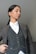 A person is wearing a formal outfit consisting of a white shirt, a dark vest with buttons and a chain detail, and a matching blazer. The hair is tied back, and the expression is neutral with a slight upward gaze.