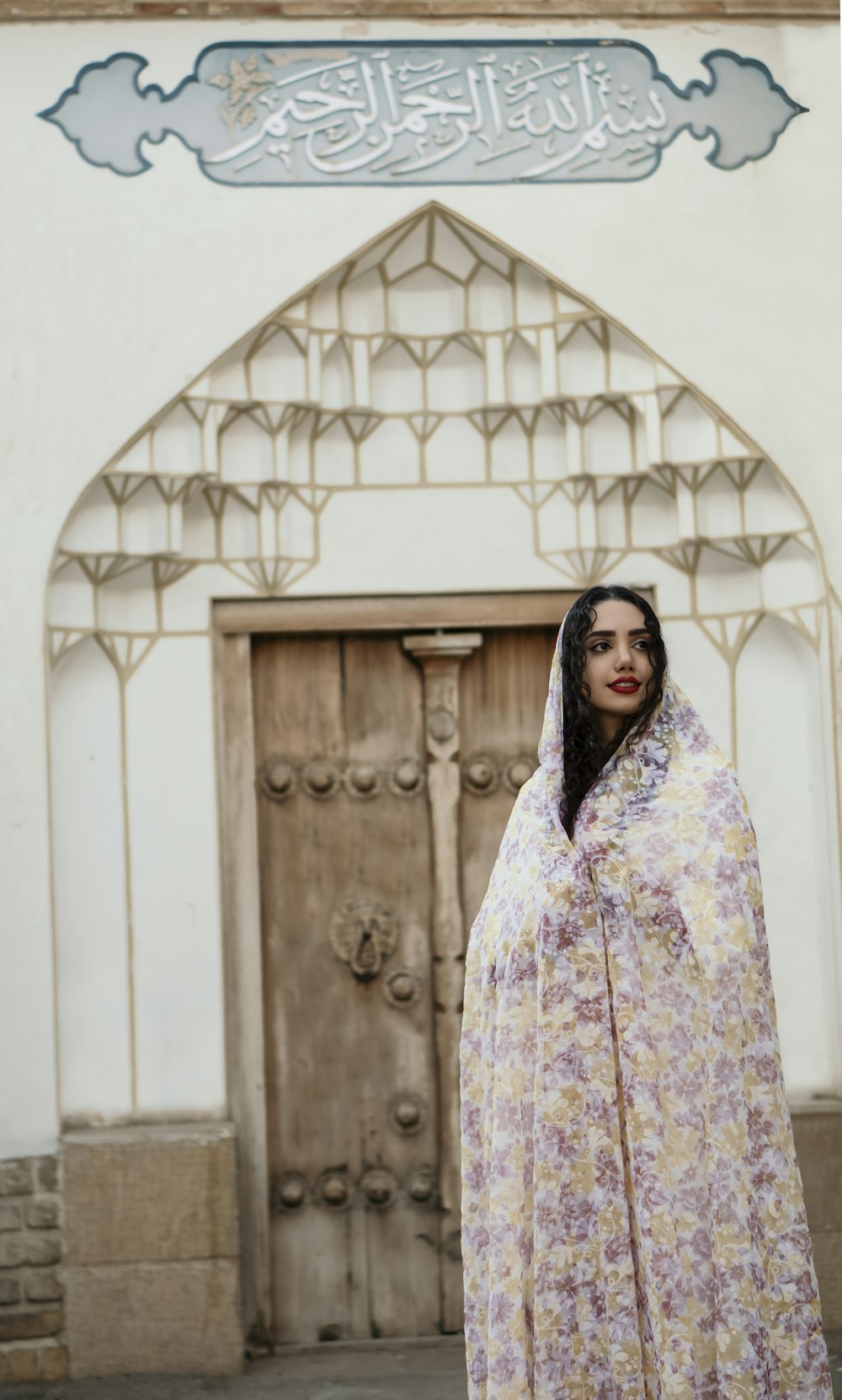 茶色の木製のドアのそばに立つ白と紫の花柄のヒジャブを着た女性