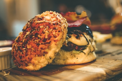 A close-up view of a gourmet hamburger sitting on a wooden surface. The hamburger is topped with melted cheese, lettuce, and crispy bacon. The bun has sesame seeds and possibly cheese baked into it. The background is softly blurred, creating a warm, inviting atmosphere.
