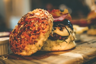 A close-up view of a gourmet hamburger sitting on a wooden surface. The hamburger is topped with melted cheese, lettuce, and crispy bacon. The bun has sesame seeds and possibly cheese baked into it. The background is softly blurred, creating a warm, inviting atmosphere.