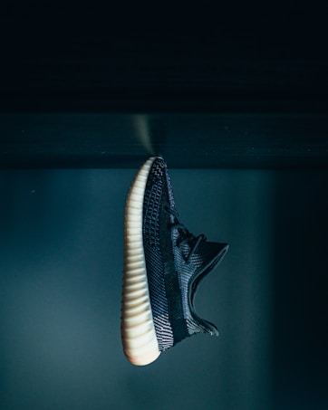 A single black sneaker is displayed against a dark, moody background. The shoe features a textured, woven pattern with a contrasting white rubber sole. The lighting highlights the intricate details of the shoe's material.