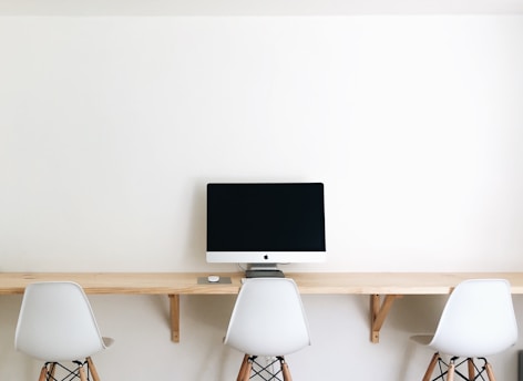 A minimalistic workspace featuring a wooden desk mounted on a white wall, with three modern white chairs. A sleek computer monitor is placed centrally on the desk.