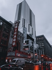 A tall, modern skyscraper stands prominently against a cloudy sky. In the foreground, the iconic red and white marquee of the Chicago Theatre is visible, with its bright lights contrasting against the reflective glass of the high-rise building. Cars and a van can be seen on the wet street below, adding a sense of urban activity. The overall setting depicts a bustling city scene.
