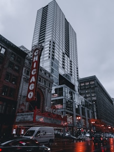 A tall, modern skyscraper stands prominently against a cloudy sky. In the foreground, the iconic red and white marquee of the Chicago Theatre is visible, with its bright lights contrasting against the reflective glass of the high-rise building. Cars and a van can be seen on the wet street below, adding a sense of urban activity. The overall setting depicts a bustling city scene.
