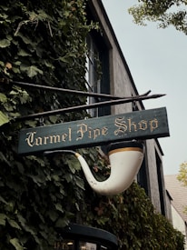 A vintage storefront sign shaped like a smoking pipe hangs on a building covered in lush green ivy. The sign reads 'Carmel Pipe Shop' in ornate lettering, and the building behind has a rustic appearance with large windows.