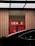 A 'No Stopping' sign is positioned in front of a glass door with red panels that display the word 'LEASE'. The scene is partially obscured by a dark vehicle, and the area is illuminated by a grid-like ceiling light.